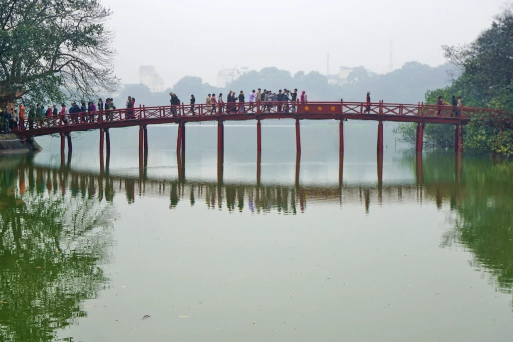 The Huc Bridge in Hanoi, Vietnam, a striking red wooden bridge leading to Ngoc Son Temple, spans across the serene Hoan Kiem Lake. This iconic landmark offers a budget-friendly cultural experience, perfect for travelers wondering if Vietnam is cheap, as entry fees are minimal. The reflection of the bridge in the calm water adds to the peaceful and picturesque setting.