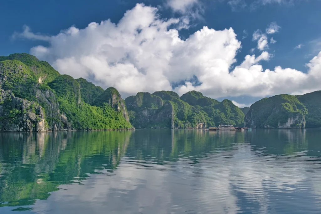 A stunning view of Ha Long Bay in Vietnam, featuring emerald green waters and towering limestone islands covered in lush vegetation, with floating houses visible in the distance. Visiting this UNESCO World Heritage site can be surprisingly affordable, making it a perfect destination for budget travelers wondering if Vietnam is cheap. The natural beauty combined with low-cost tours offers exceptional value for money.