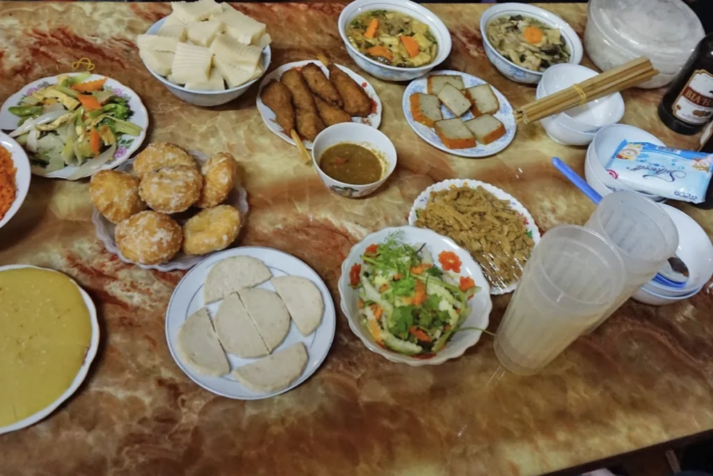 A table filled with traditional Vietnamese dishes, including a plate of fried doughnuts coated in sugar, sliced steamed rice cakes, a bowl of bamboo shoots, fried fish skewers, a bowl of dipping sauce, and plates of mixed vegetables and pickled vegetables. Other dishes include sliced sausage, stir-fried mushrooms with carrots, and a bowl of shredded chicken or pork. Disposable plastic cups, chopsticks, and a bottle of beer are also visible on the table.