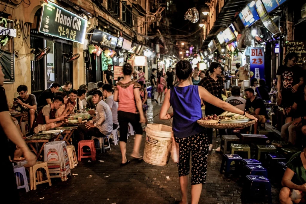 Crowded Hanoi night market with locals and tourists enjoying street food and shopping, while a woman carries a basket and pail among small tables and food stalls.
