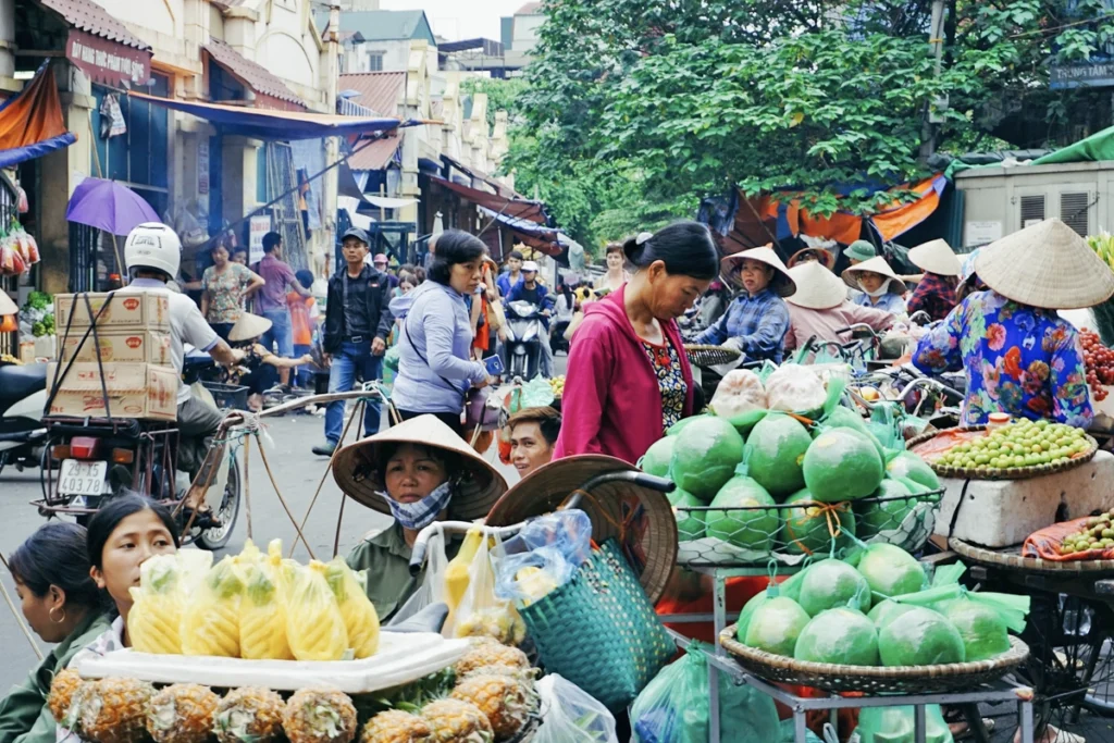 
A bustling street market in Hanoi, Vietnam, filled with fresh produce like peeled pineapples, pomelos, and various fruits sold from baskets and bicycles. Local vendors wearing traditional conical hats offer affordable food options, showcasing why Vietnam is affordable for travelers seeking fresh, budget-friendly meals. The vibrant atmosphere highlights the culture of low-cost street markets in Vietnam.