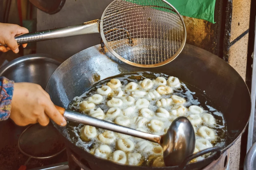 A vendor frying traditional Vietnamese dumplings in a large wok filled with hot oil, using a metal ladle and skimmer to cook the golden, doughy treats. The bubbling oil and stainless steel kitchen tools suggest a street food preparation scene.