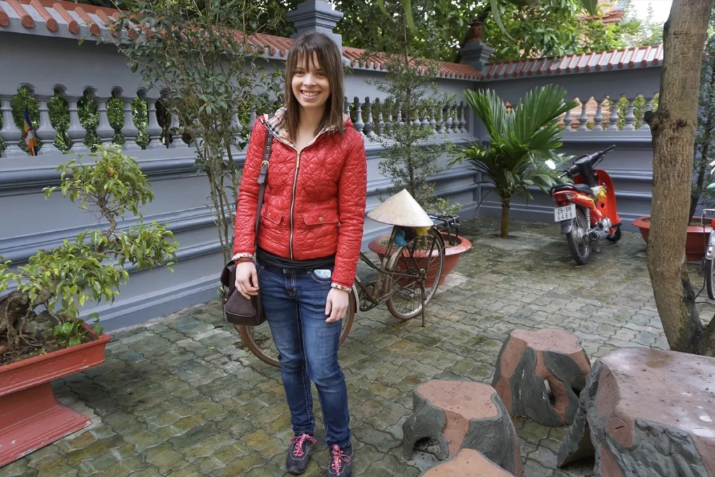 A person in a red jacket and jeans smiling in a peaceful courtyard in Hanoi, Vietnam, with a traditional bicycle, a conical hat, and a red motorbike in the background. The courtyard is surrounded by gray walls with intricate detailing, potted plants, and stone stools. The setting reflects the blend of modern and traditional elements commonly seen in Vietnam.