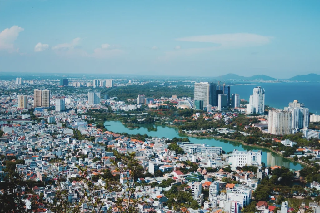 A panoramic cityscape of a coastal city in Vietnam, featuring a mix of modern high-rise buildings and traditional low-rise homes. The view also captures a lake surrounded by urban development, showing the blend of modern growth and cultural heritage in Vietnam.