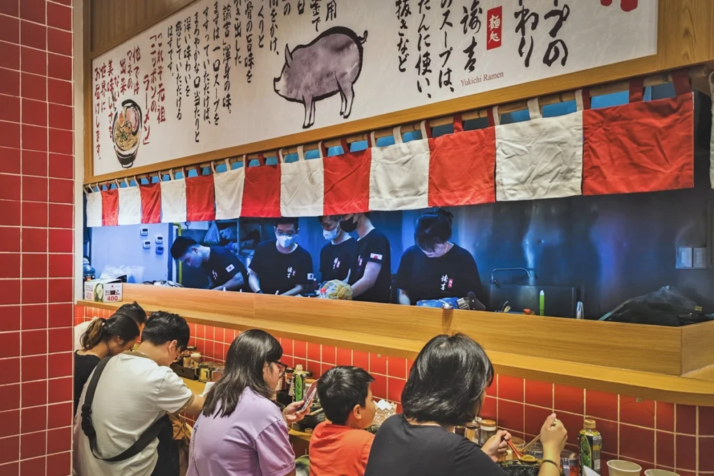 
A cozy ramen restaurant in Vietnam with customers enjoying bowls of noodles at a counter while chefs prepare meals behind a half-curtain, emphasizing the affordable dining experience the country offers. The red and white decor and Japanese-style illustrations on the wall add to the authentic atmosphere, showcasing how dining out in Vietnam can be budget-friendly while still offering quality food. The price tag of "400" on the counter further suggests how cheap meals can be compared to Western standards.