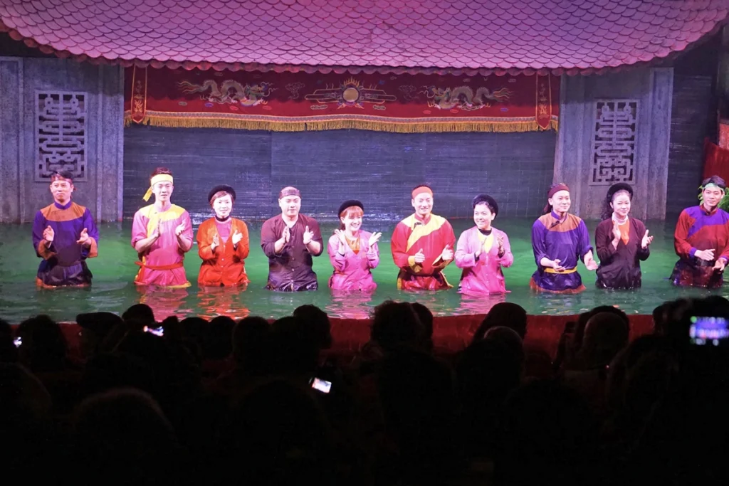 A traditional Vietnamese water puppet performance featuring performers in colorful costumes standing waist-deep in water, applauding the audience. The ornate stage with dragon motifs and a red curtain highlights the cultural significance of this art form. This unique experience offers an affordable way for travelers to explore Vietnam's rich cultural heritage.