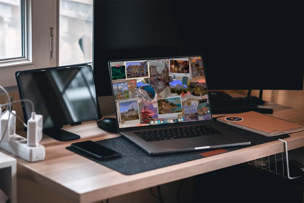 A MacBook laptop on a modern desk displaying a vibrant travel vision board filled with photos of famous landmarks, nature scenes, and cultural sites. The workspace also features a smartphone, notebook, secondary monitor, and a power strip, with natural light streaming through a window.