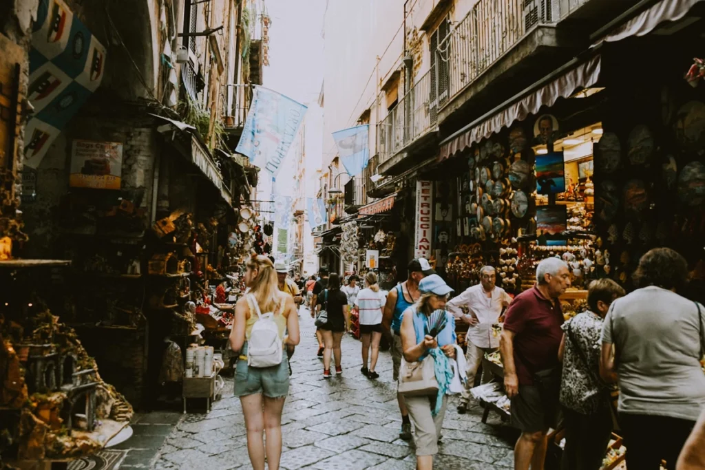 A bustling narrow street market in Italy filled with tourists and locals shopping for souvenirs, crafts, and artwork. The vibrant market scene captures the charm of exploring Italian culture.