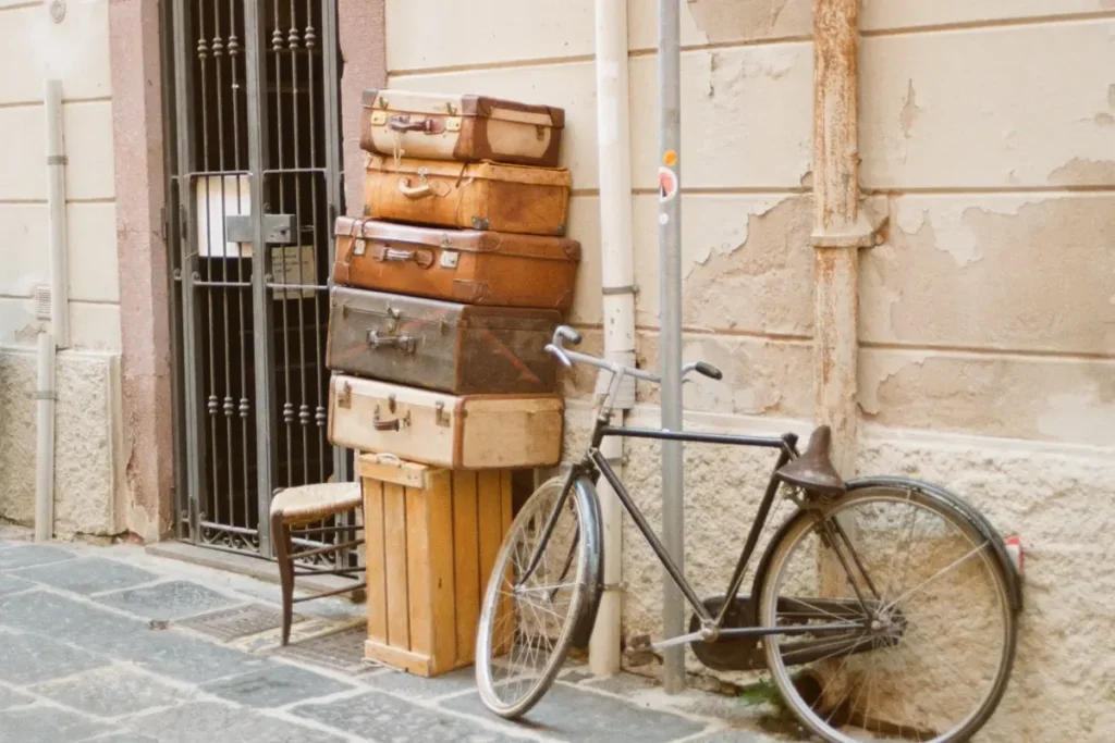 A stack of vintage suitcases next to an old bicycle against a weathered Italian wall. The retro travel theme emphasizes packing stylish yet practical luggage for a European trip.